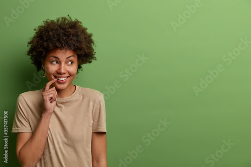 Portrait of optimistic glad curly woman looks away with engaging toothy smile, expresses joy and happiness, wears casual beige t shirt, isolated over green background, copy space for promo or text