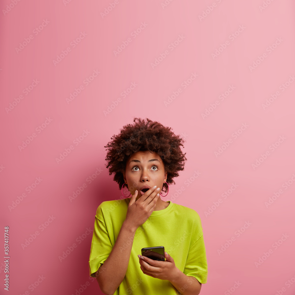 Astonished dark skinned woman expresses amazement as reads message, gasps from wonder, covers mouth and looks upwards, uses mobile phone, isolated over rosy wall, blank space for your promo. Astonished dark skinned woman expresses amazement as reads message, gasps from wonder, covers mouth and looks upwards, uses mobile phone, isolated over rosy wall, blank space for your promo.