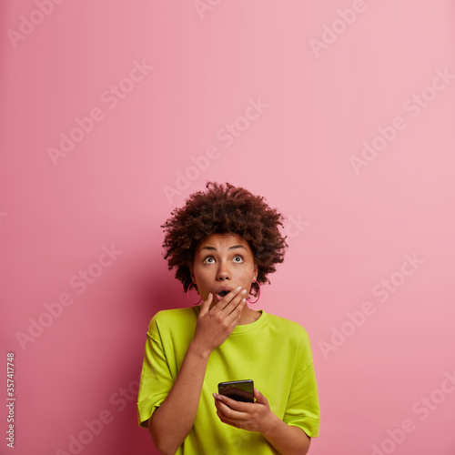 Astonished dark skinned woman expresses amazement as reads message  gasps from wonder  covers mouth and looks upwards  uses mobile phone  isolated over rosy wall  blank space for your promo.