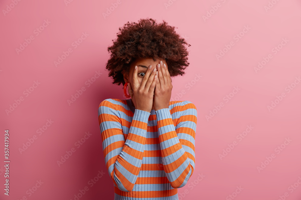 Scared ethnic woman peeps through fingers, hides face with hands, looks terrified, afraids of something terrible, wears casual striped jumper, isolated on pink background. People and phobia. Scared ethnic woman peeps through fingers, hides face with hands, looks terrified, afraids of something terrible, wears casual striped jumper, isolated on pink background. People and phobia.