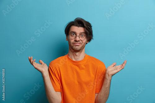 Clueless adult man says dont know, maybe, who cares, looks disinterested in question, has doubts, stands bewildered, spreads palms sideways, wears optical glasses and orange t shirt, isolated on blue photo