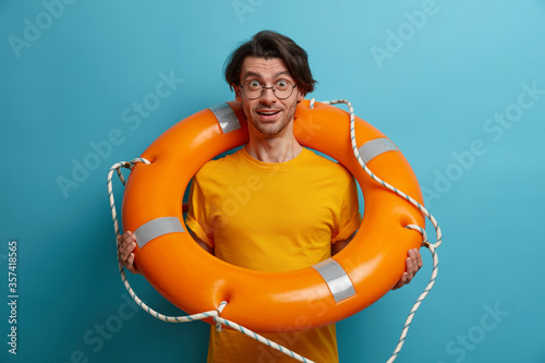 Pleased cheerful man swimmer stands with lifebuoy, listens instructions from lifesaver before swimming in sea, wears glasses and yellow t shirt, enjoys summer vacation. People and recreation
