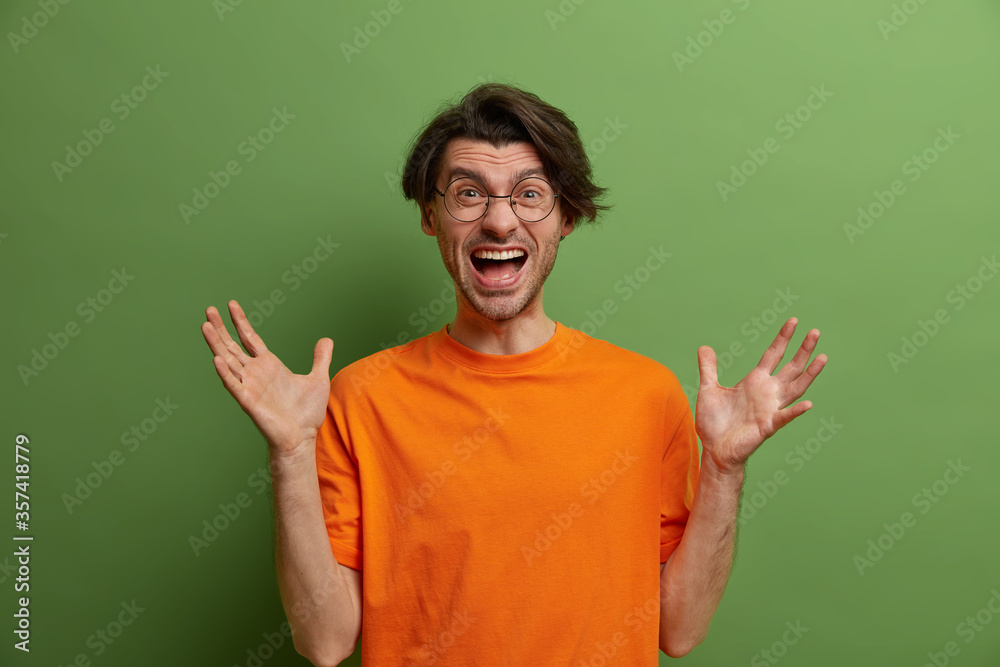 Angry disappointed man reacts emotionally on negative news, exclaims loudly, raises hands, screams with annoyance, cannot control his emotions, wears spectacles and orange t shirt, poses indoor Angry disappointed man reacts emotionally on negative news, exclaims loudly, raises hands, screams with annoyance, cannot control his emotions, wears spectacles and orange t shirt, poses indoor
