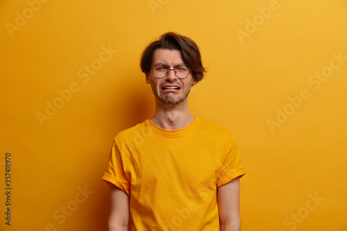 Distressed man with sorrowful expression, cries with despiar, regrets doing something wrong, has big problems, squints face, wears optical glasses, dressed in yellow t shirt. Negative emotions