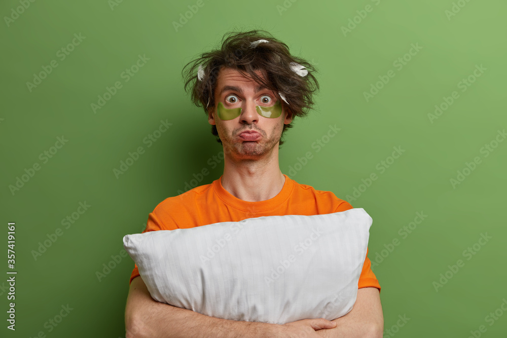 Fototapeta premium Displeased sullen man embraces pillow, looks with unhappy sleepy expression, wakes up early to have beauty treatments, wears under eye pads for removing wrinkles and fine lines, cares about complexion
