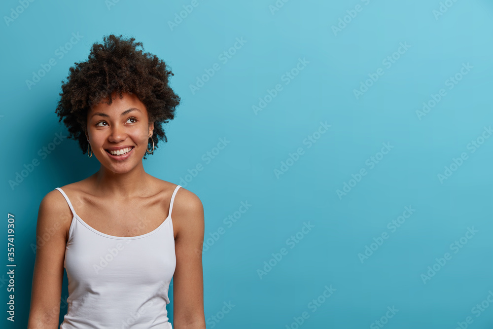 Smiling positive young woman with Afro hair looks away with dreamy expression, thinks about good future, has toothy smile, wears casual t shirt, poses against blue background, empty space for text Smiling positive young woman with Afro hair looks away with dreamy expression, thinks about good future, has toothy smile, wears casual t shirt, poses against blue background, empty space for text