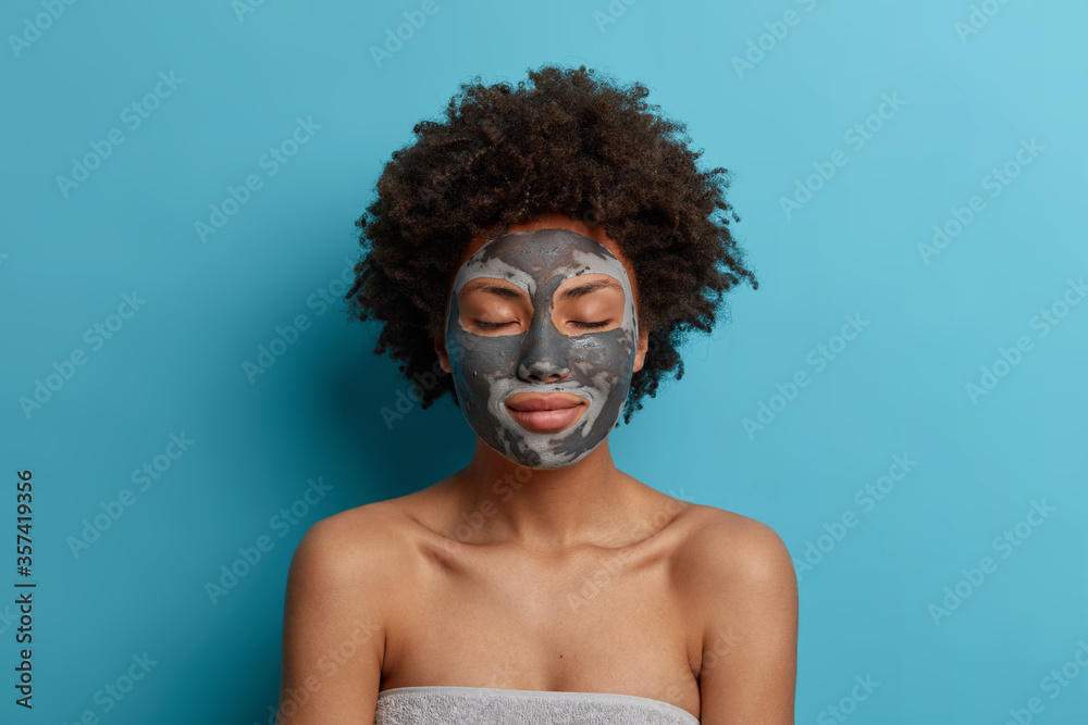 Beautiful young woman has clean fresh skin, wears beauty clay mask on face, stands with closed eyes, cares about body after taking shower, enjoys beauty treatments, stands with bare shoulders