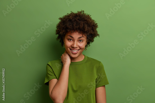 Studio shot of beautiful woman with perfect skin and toothy smile, touches neck, being in good mood, has casual talk, wears summer green t shirt, poses indoor. Positive human emotions concept