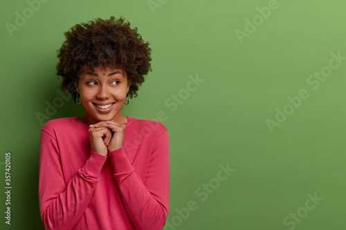 Good looking pleased ethnic curly woman has good mood and expresses positive emotions, keeps hands under chin, looks aside, has toothy smile, dressed in rosy jumper, isolated on green background