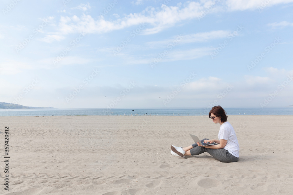 Smiling pretty woman sitting on the beach with her laptop. Freelance concept.