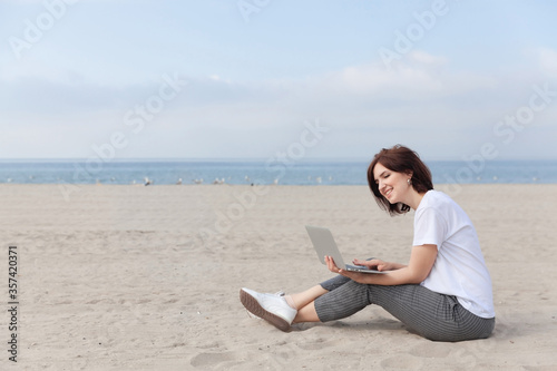Smiling pretty woman sitting on the beach with her laptop. Freelance concept. © Iryna Dincer