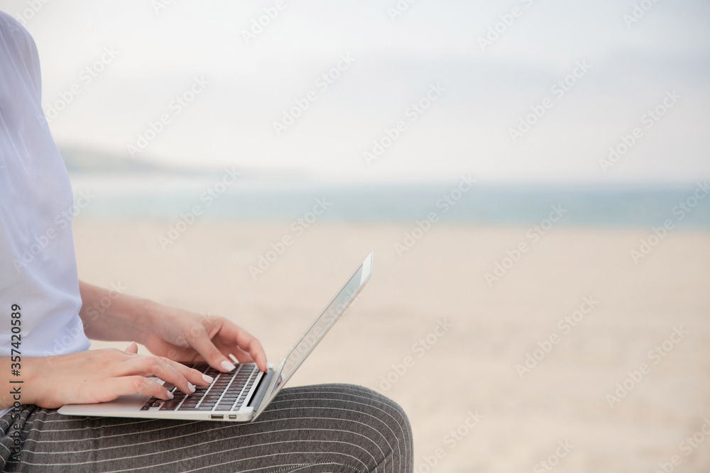 Close up of woman's hands typing on laptop with beach and sea on the background.