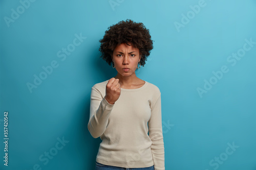 Outraged furious woman with Afro hair, clenches fist with anger, experiences strong aggression, smirks face, wears casual white long sleeve jumper, expresses irritation, stands against blue wall