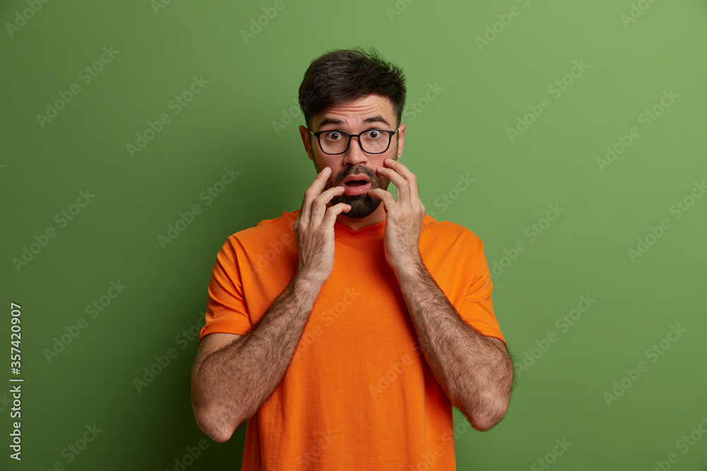 Scared nervous hipster grabs face, looks frightened at camera, keeps mouth opened, cannot believe eyes, wears casual orange t shirt, poses against green background. People and emotions concept