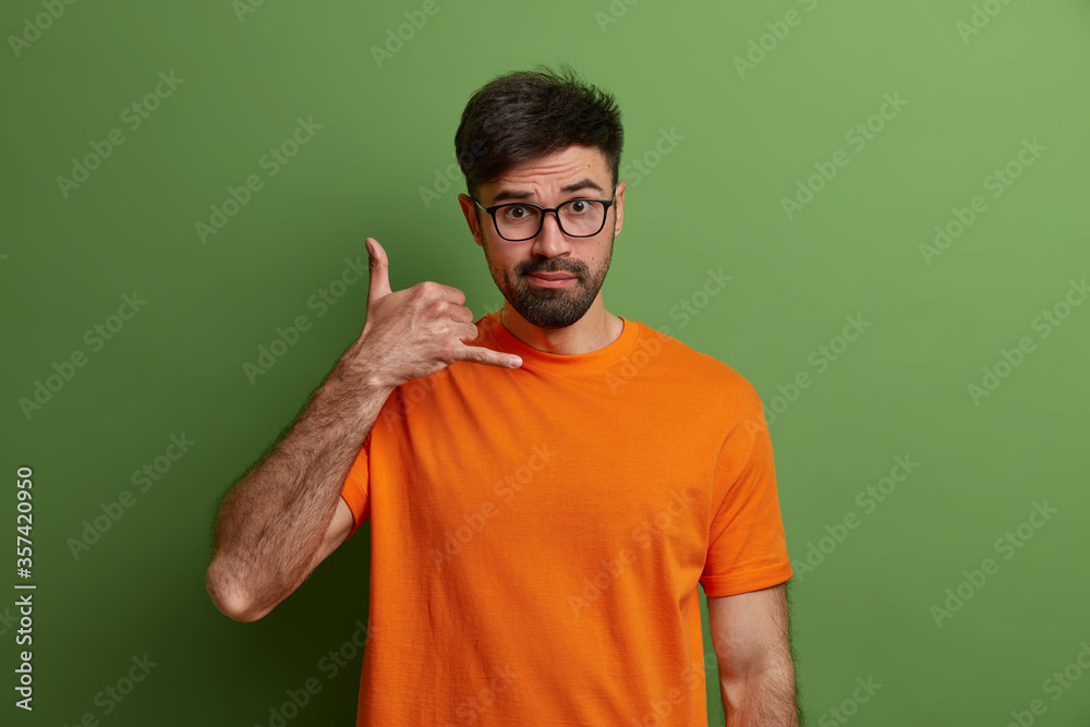 Lets contact by phone. Serious unshaven man makes phone gesture, shows call me hand sign, waits for call, wears orange t shirt, transparent eyeglasses, isolated on green wall. Body language concept Lets contact by phone. Serious unshaven man makes phone gesture, shows call me hand sign, waits for call, wears orange t shirt, transparent eyeglasses, isolated on green wall. Body language concept
