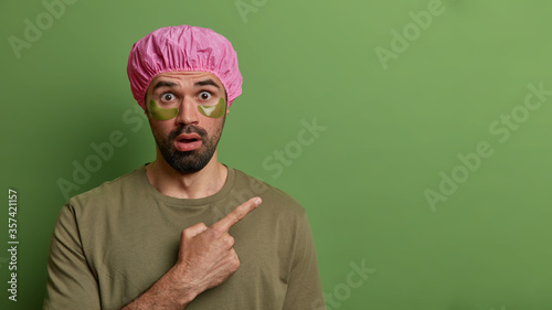 Horizontal view of shocked bearded man points index finger on empty space, has bugged eyes, applies collagen patches for reducing wrinkles or puffiness, isolated on green background takes care of skin
