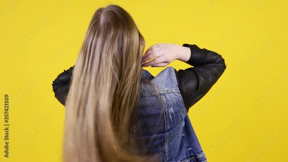 Beautiful young woman wearing a denim jacket with long wavy hair on yellow background. Back view.