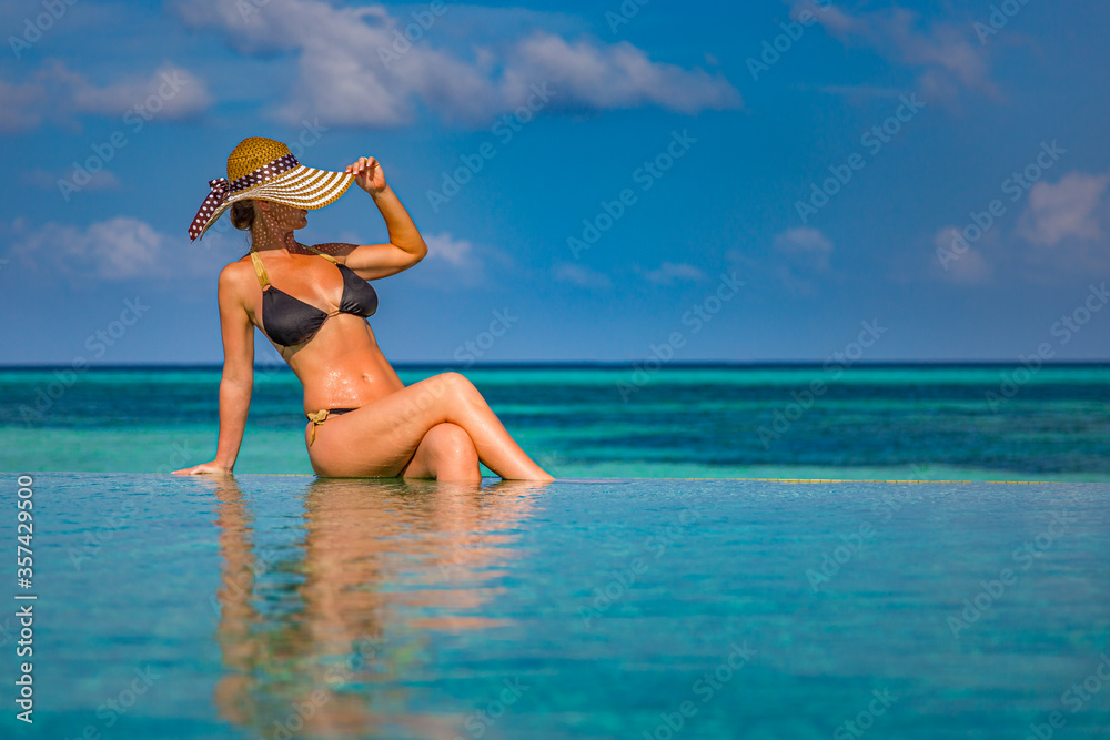 Young woman in bikini enjoying a sun. Luxury tropical vacation and summer holiday concept. Woman relaxing in infinity swimming pool looking at view. Luxury summer beach background Young woman in bikini enjoying a sun. Luxury tropical vacation and summer holiday concept. Woman relaxing in infinity swimming pool looking at view. Luxury summer beach background