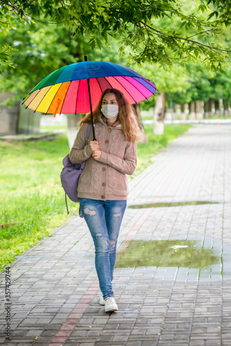 Caucasian girl in a protective mask walks under an umbrella on an empty street in spring rain. Safety and social distance during a coronavirus pandemic. New normal, the implications of quarantine