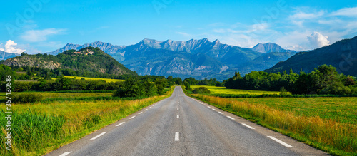 A long straight road leading towards a mountains in France. Amazing bright colorful spring and summer landscape. Yellow fields of flowering rape and blue sky with clouds. 
