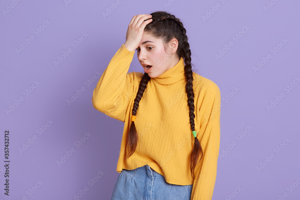 Girl with long dark hair and pigtails standing with opened mouth, keeping hand on forehead, forget something important, woman standing against lilac wall. Girl with long dark hair and pigtails standing with opened mouth, keeping hand on forehead, forget something important, woman standing against lilac wall.
