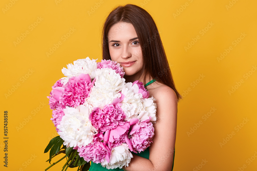 Romantic girl embracing huge bouquet of flowers in hands, looking directly at camera, enjoying beautiful smell of flowers, stands against yellow wall. Romantic girl embracing huge bouquet of flowers in hands, looking directly at camera, enjoying beautiful smell of flowers, stands against yellow wall.