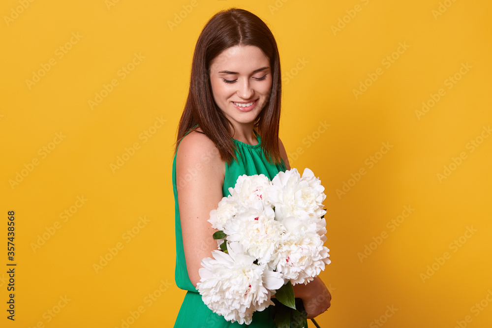 Brunette woman with charming smile wearing green dress, holding bouquet of white peonies and looking at flowers with gentle, standing isolated over yellow background. Brunette woman with charming smile wearing green dress, holding bouquet of white peonies and looking at flowers with gentle, standing isolated over yellow background.