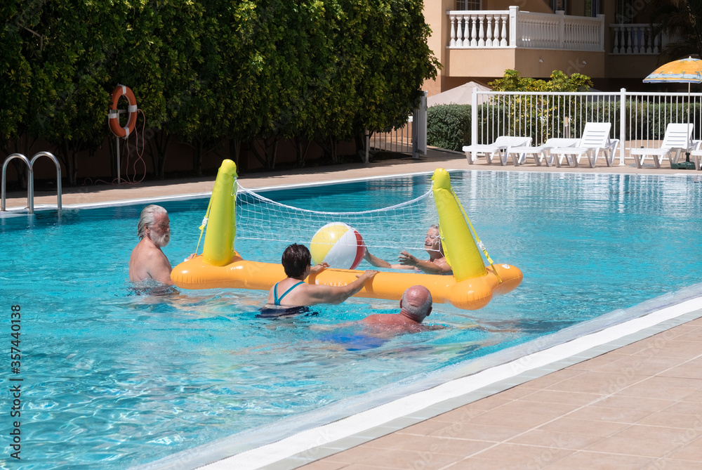 Four senior people friends or family in active elderly vacation having fun in swimming pool with balloon and volleyball - happy retirement concept under the summer sun