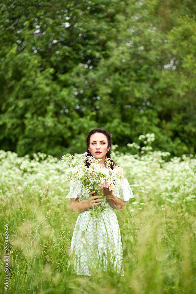 beautiful young brunette woman in a green dress on the meadow with white flowers in a hand on a warm summer day beautiful young brunette woman in a green dress on the meadow with white flowers in a hand on a warm summer day