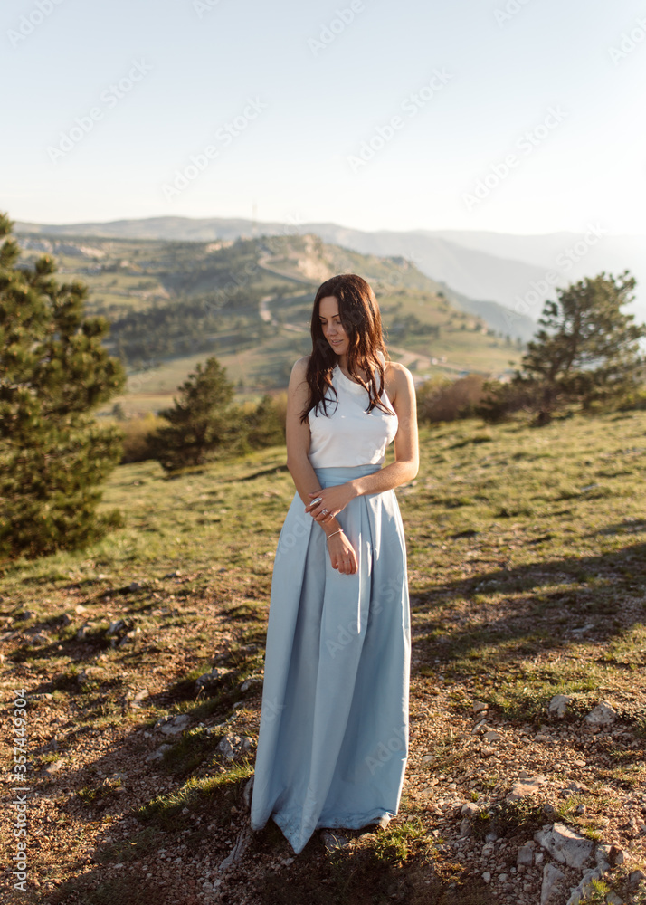Beautiful bride in white blouse and blue skirt standing on the picturesque hill in the sunlight. An attractive girl in a minimalistic dress with jewelry rings on her hands stands in a green field.