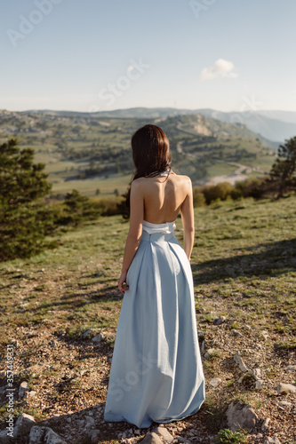 A bride with a slim figure in blue dress with an open back stands on a green field against hills.