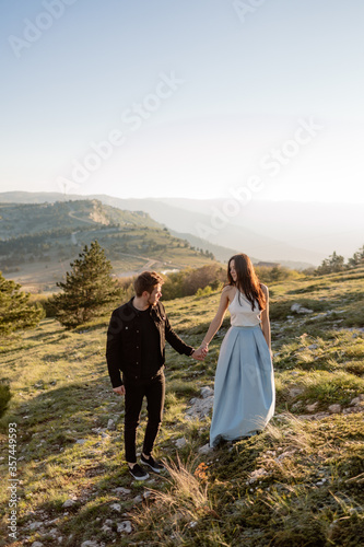 Bride and Groom walk hand in hand across picturesque hill with green flowering grass and spruce trees in mist light. A bride in a blue dress holds the hand of her stylish groom in black suit