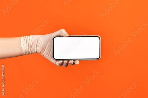 Profile side view closeup of human hand in white surgical gloves holding and showing horizontal smart phone empty display. indoor, studio shot, isolated on orange background.