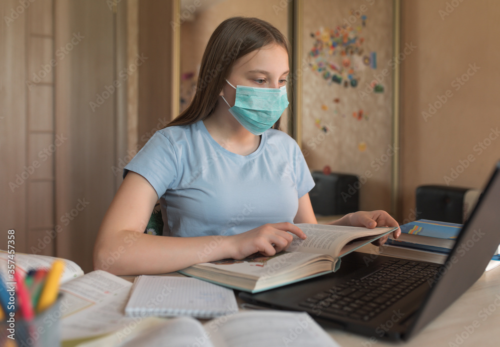 Teenager girl, in a medical mask, disinfection from bacteria and germs and viruses, reads a paper book, laptop monitor, e-education and online lessons, distance learning.