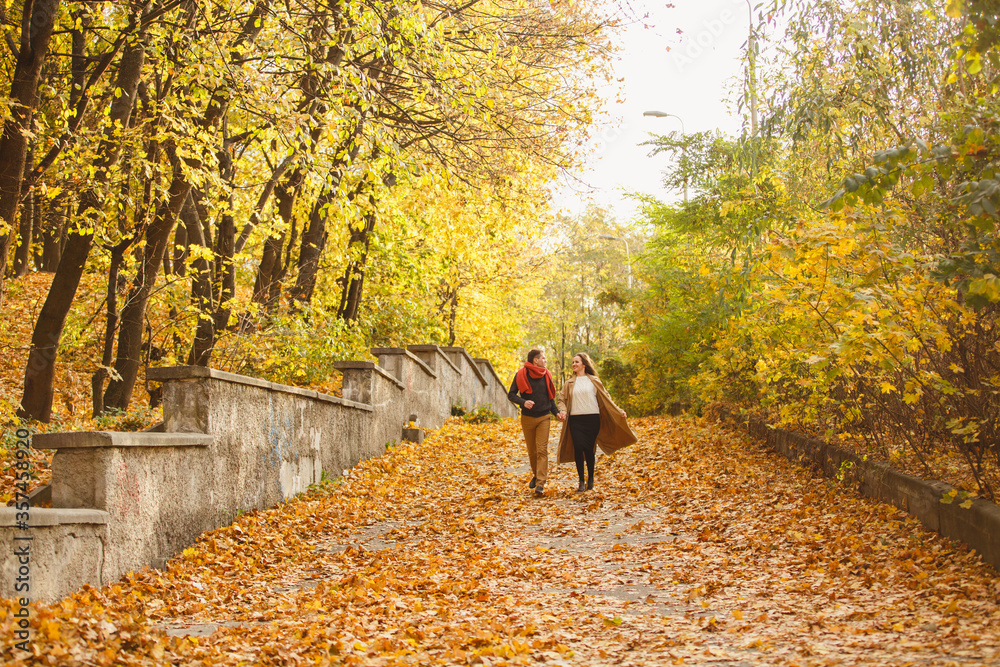 Couple weared in autumn-style clothes (oranfe scarf and vest) running through the autumn landscape. Alley covered with yellow foliage. Autumn walk outdoors. Two lovers in autumn park. Romantic dating