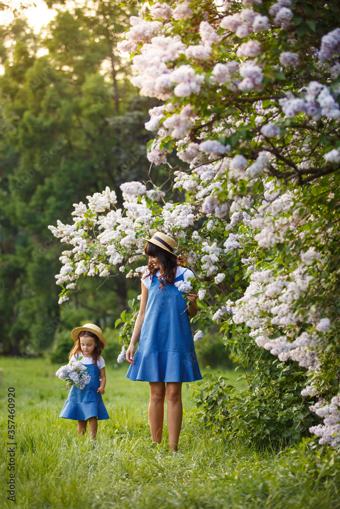 Fototapeta premium Mother and little daughter on picnic in blooming lilac garden. Mom loves her child. Spring story. Family look romantic similar blue dresses and straw hat. Happy family in beautiful spring day