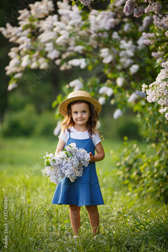Little girl weared in romantic rustic look in a blooming lilac garden with lilac bouquet. Spring story. Romantic rustic look with blue dress and straw hat. Happy girl in beautiful spring day. 