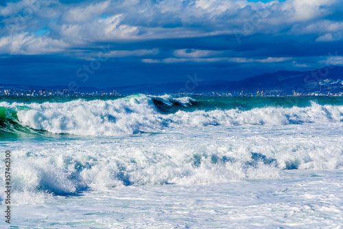 Storm waves in white foam rush in rows along the Tsemesskaya Bay. Blue sky and green sea. Dangerous and dramatic. In the background, mountains, multi-storey buildings of the city and port. © Александр Трихонюк