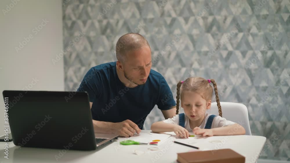 shaved bald father helps glue colorful sticker on paper sheet to pretty little daughter sitting at white table with laptop in room at isolation shaved bald father helps glue colorful sticker on paper sheet to pretty little daughter sitting at white table with laptop in room at isolation