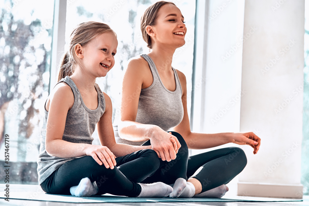 Young mother sitting with her 3-years daughter in lotus pose asana on window sill in front of window Young mother sitting with her 3-years daughter in lotus pose asana on window sill in front of window