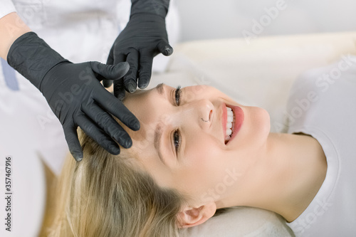 Cropped image of hands of female professional cosmetologist making face massage to young smiling woman feeling relaxed in beauty salon. Concept of antiaging facial skin treatments and massage