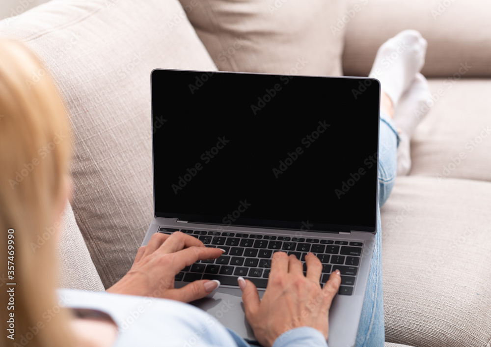 Fototapeta premium Work online. Woman typing on laptop with blank screen, lying on sofa in home interior, shoulder view