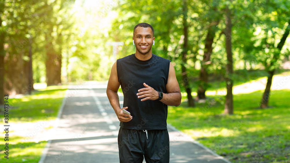 Smiling black guy with mobile device and earphones jogging at park on sunny day Smiling black guy with mobile device and earphones jogging at park on sunny day