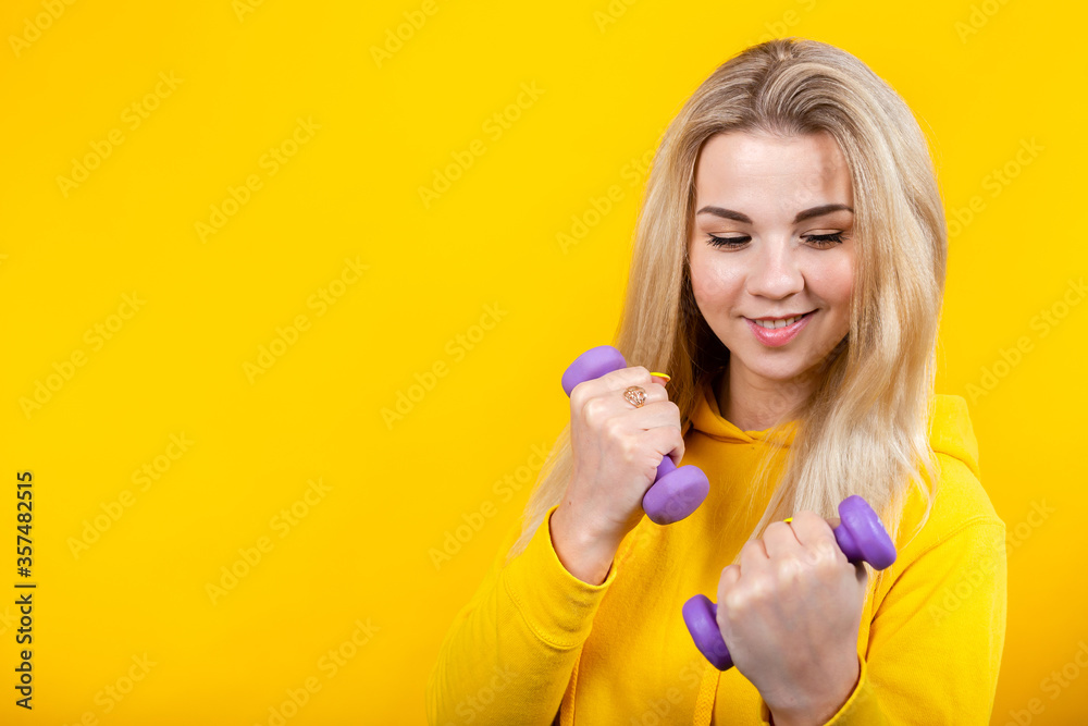 Attractive beautiful young blonde woman in casual yellow sporty clothes doing exercises with small 0.5 kg purple dumbbells. Attractive beautiful young blonde woman in casual yellow sporty clothes doing exercises with small 0.5 kg purple dumbbells.