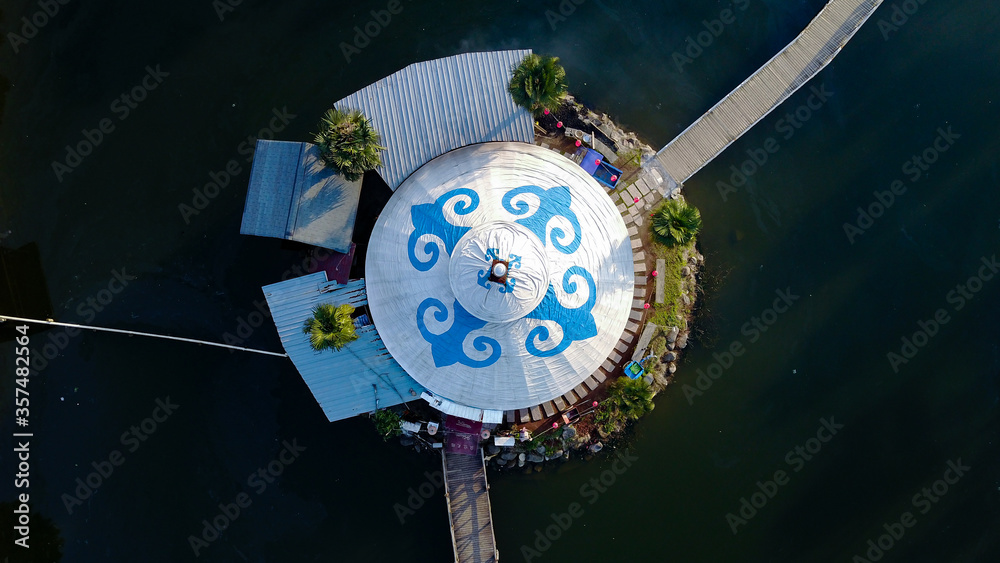 Aerial view on Mongolian white styled Yurt with blue ornaments in the middle of a small water reservoir with two bridges. 