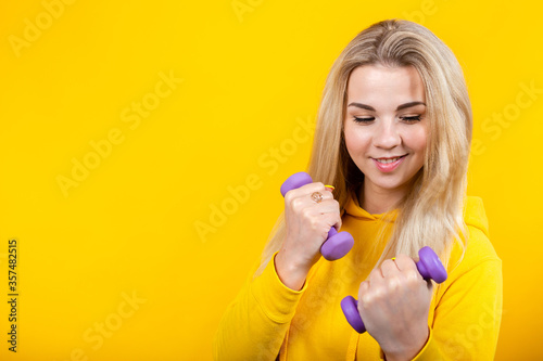 Attractive beautiful young blonde woman in casual yellow sporty clothes doing exercises with small 0.5 kg purple dumbbells.