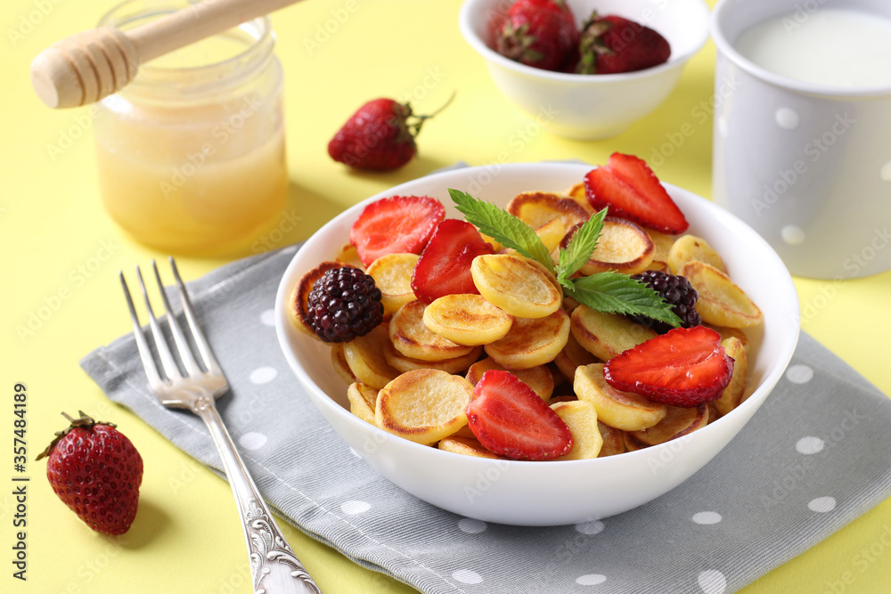 Trendy tiny pancakes for breakfast with strawberry, blackberry and honey in white bowl on yellow background. Closeup, Horizontal format Trendy tiny pancakes for breakfast with strawberry, blackberry and honey in white bowl on yellow background. Closeup, Horizontal format