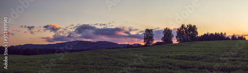 panorama landscape at sunset - meadow, trees and hills and clouds illuminated by the setting sun