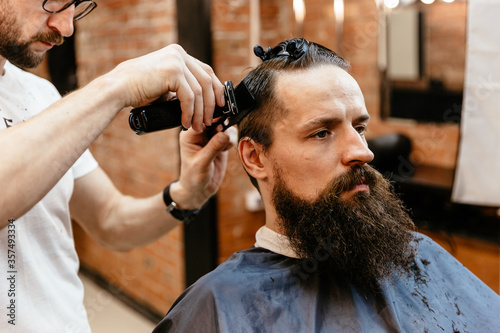 Grooming of real man. Side view of young bearded man getting beard haircut at hairdresser while sitting in chair at barbershop