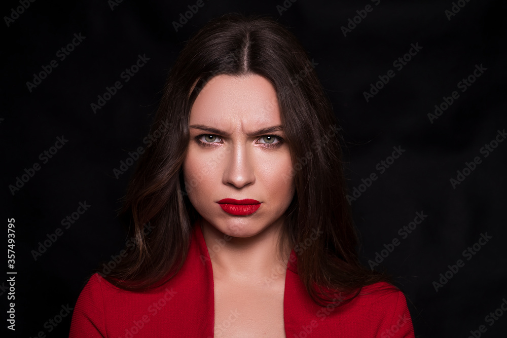 Emotional head shot portrait of a brunette caucasian woman in red dress and with red lips on black background. She make sad emotion